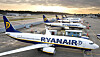 Flera Ryanair-plan parkerade på en flygplats med soluppgång och terminaler i bakgrunden.