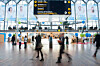 Blurred passengers walk through a bright terminal beneath a large electronic information board.