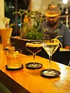 Kolme cocktailia puupöydällä, taustalla epäselvä baarimikko ja yrttiruukku.