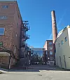 Tiilirakenteinen tehdaspiha, jonka keskellä lasinen sisäänkäynti ja korkea savupiippu.