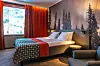Hotellihuone, jossa metsäaiheinen seinäkuva, parivuode ja oranssit verhot ikkunan edessä.