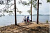 Kaksi pyöräilijää seisoo polkupyörien kanssa järven rannalla puiden välissä.