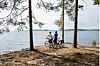 Kaksi pyöräilijää seisoo polkupyörien kanssa järven rannalla puiden välissä.