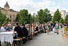 Ihmiset istuvat pitkien pöytien ääressä ulkona kävelykadulla rakennusten ja puiden ympäröimänä.