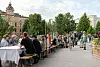 Ihmiset istuvat pitkien pöytien ääressä ulkona kävelykadulla rakennusten ja puiden ympäröimänä.