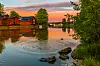 Joki, kivinen ranta, puutaloja ja puiden reunustama maisema auringonlaskussa.