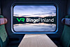 Junavaunun sisätila, jossa pöytä ja istuimet sekä ikkunanäytöllä VR Binge Finland -logo ja maisema.