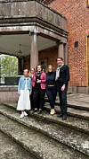 Neljä ihmistä seisoo portaitten yläosassa punatiilisen rakennuksen sisäänkäynnin edessä.