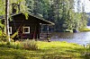 Pieni hirsimökki järven rannalla metsäisessä maisemassa.