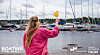 Nainen seisoo venemessuilla ja osoittaa näyttöä satamalaitureiden ja purjeveneiden edessä.
