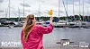 Nainen seisoo venemessuilla ja osoittaa näyttöä satamalaitureiden ja purjeveneiden edessä.