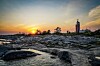 Kivinen rantakallio, matala vesi ja majakka auringonlaskussa.