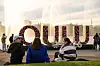 Suuri Oulu-tekstitaideteos aukiolla, ja muutama ihminen seisoo sen edessä iltavalossa.