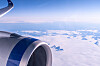 Vy från ett flygplansfönster med en stor jetmotor och blå himmel ovanför vita moln.