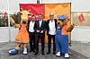 Three men stand between two airline mascots in orange and blue outfits at a branded event backdrop.