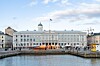 Vaalea rantarakennus satama-altaalla, oranssi vene laituriin kiinnitettynä ja ihmisiä kävelemässä rannassa.