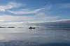 Pieni vene ja veneessä seisova henkilö tyynellä merellä avaran taivaan alla.