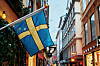 En svensk flagga hänger på en gata i Gamla stan, Stockholm med butiksfönster och byggnader i varmt ljus.