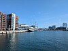 Vesistö, modernit asuintalot, venesatama, purjeveneitä ja silta kirkkaan taivaan alla.