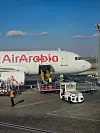 Air Arabian matkustajakone ja maahuoltoajoneuvoja lentokentän asematasolla sinisen taivaan alla.