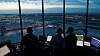Silhuetter av flygledare vid datorskärmar med utsikt över en flygplats.