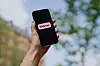 En hand håller upp en svart mobiltelefon med Klarnas rosa logotyp mot en suddig utomhusbakgrund.