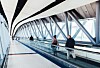 Resenärer åker på ett rullband i en ljus flygplatsterminal med glasväggar och stålkonstruktioner.