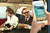 En smartphone med texten ”Find your wave” hålls framför två resenärer som äter i en äldre flygplanskabin.