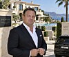 Kim Fournais in a dark suit outside a villa with palm trees, hills and a black car.