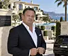 Kim Fournais in a dark suit outside a villa with palm trees, hills and a black car.