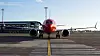 Rött och vitt passagerarflygplan sett framifrån på en flygplats med terminal och blå himmel i bakgrunden.