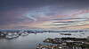 Panoramavy över en hamn med fartyg, småbåtar och stadsbebyggelse under en färgstark kvällshimmel.