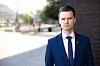 Tummansiniseen pukuun pukeutunut mies seisoo tiiliseinän vieressä kaupunkikadulla.